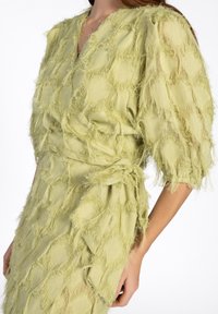 Hellgrünes Kleid mit strukturiertem, fransigem Finish, das über einen V-Ausschnitt und eine gebundene Taille verfügt. Kurze, puffige Ärmel verleihen dem Kleid mehr Dimension.