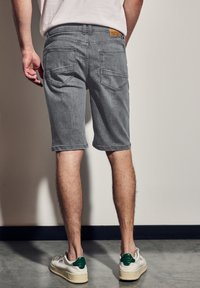 Harmaat farkkushortsit suoran leikkauksen omaavina, joissa on kaksi takataskua ja vaaleanruskea etiketti vyötäröllä. Yhdistettynä valkoisiin tennareihin, joissa on vihreitä yksityiskohtia.