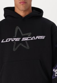 Sort hættetrøje lavet af blødt stof med et design af rhinestones i form af en stjerne og teksten "LOVE SCARS" i lilla. Indeholder en frontlomme og hætte.