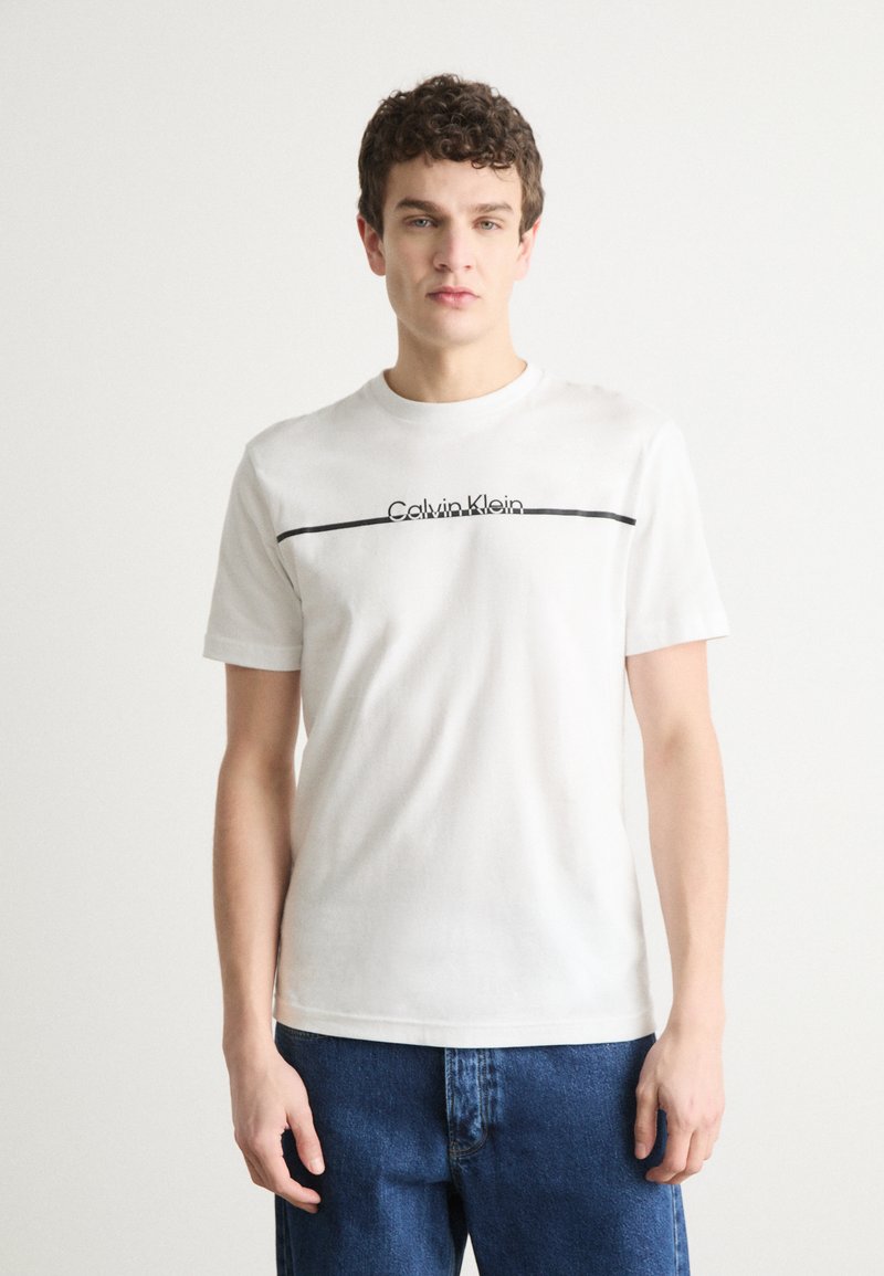 Hvid t-shirt i bomuld med et sort Calvin Klein-logo og vandret linje på brystet. Korte ærmer, rund halsudskæring, afslappet pasform.