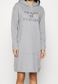 Vestido sudadera gris hecho de tela suave, con un bolsillo frontal y capucha con cordón. Logo "TOMMY HILFIGER" en coloridos strass.