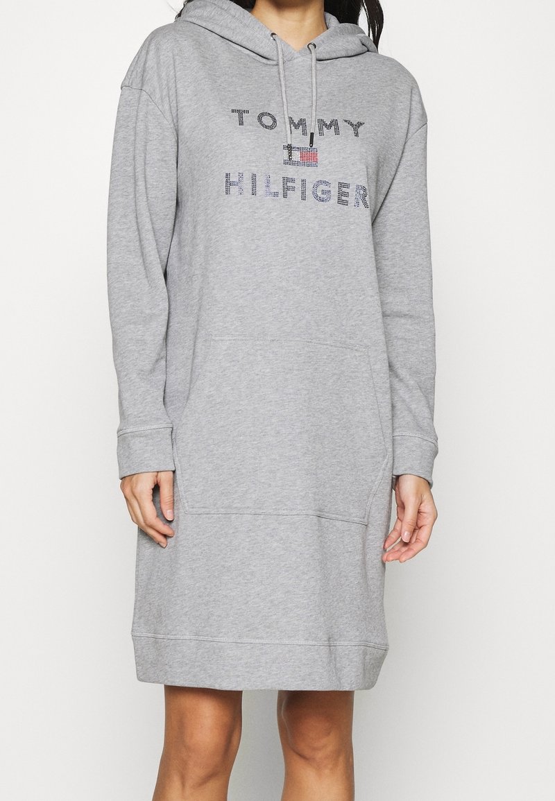 Vestido sudadera gris hecho de tela suave, con un bolsillo frontal y capucha con cordón. Logo "TOMMY HILFIGER" en coloridos strass.