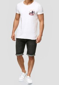 Bílý tričko s krátkým rukávem, které má design kapes s palmou v růžové a černé barvě, v kombinaci s tmavými džíny s ohrnutými nohavicemi a bílými teniskami.