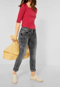 Mörkgrå denimjeans med tapered fit och uppvikta muddar, kombinerat med en fuchsian färgad långärmad topp och beige sneakers. En gul tröja hängande över armen.