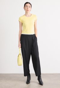 Haut jaune à manches courtes avec des bordures blanches, associé à un pantalon noir ample. Le mannequin tient un petit sac à main jaune et porte des bottines noires.