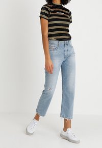 Camiseta de manga corta a rayas negras y amarillas con el texto "BAD THINGS", combinada con jeans rectos descoloridos de color azul claro y zapatillas blancas.