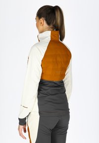 Tætsiddende jakke i creme og mørkegrå med en puffy orange skulderdel; har høj krave og elastiske ærmer. Matchet med strækbare grå bukser.
