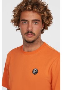 Orange bomulds T-shirt med korte ærmer, rund hals, med et sort rundt logo-patch på brystet. Glat tekstur og enkelt design.