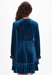 Mélykék bársony ruha hosszú ujjakkal, összehúzott derékkal és fodros szegéllyel. Sima textúra, lágy fényű.