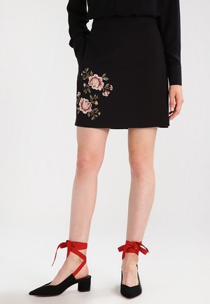 Sort mininederdel med pink blomsterbroderi båret med sorte høje hæle med røde ankelsløjfer på en stående person.