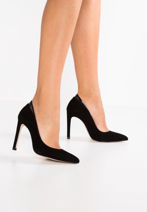 Sorte ruskinds-pumps med høje hæle, spids tå, glatte sorte lakdetaljer og en slank stilet-hæl, med et lavtudskåret design.