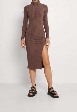 Vrouw draagt een bruin ribgebreide midi-jurk met col en een hoge split aan de dij, gecombineerd met zwarte platte sandalen met enkelbandjes.
