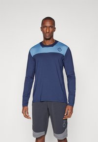 Marineblauw sportshirt met lange mouwen en blauwe accenten, ronde hals, vochtafvoerende stof, gecombineerd met grijze shorts met een subtiel logo en ontwerp.