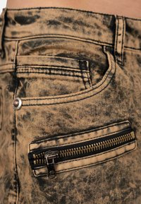 Jeans in denim marrone con effetto usurato e dettagli neri, dotati di tasca con zip e cuciture in vista lungo i bordi. Motivo di superficie testurizzato.