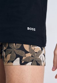 Melns T-krekls ar balto "BOSS" logo, kombinēts ar rakstainām šortiem ar bēša un melnā krāsā lapu motīviem. Gluda auduma, pieguloša forma.