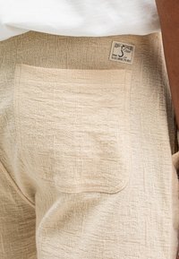 Beige strukturierte Shorts mit einer großen Rückentasche und einem gewebten Muster. Enthält ein Etikett mit der Aufschrift "BUTCHER" am Bund.