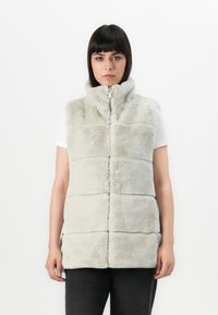 Grijze nepbont vest met hoge kraag, rits aan de voorkant en gequiltte panelen. Ggedragen over een wit shirt met een zachte, gestructureerde oppervlakte.