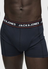 Gros plan d'une personne portant un boxer bleu marine Jack & Jones avec le logo visible sur la ceinture, sur une peau foncée.
