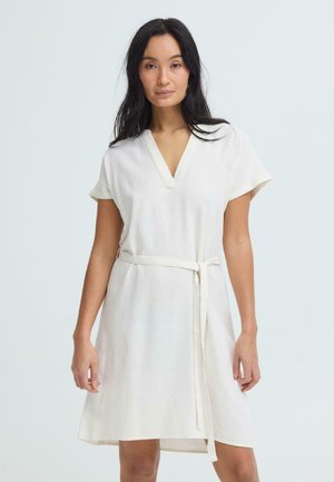 Woman with long dark hair wearing a short-sleeved white dress with a V-neck and a waist tie, standing against a plain light background.