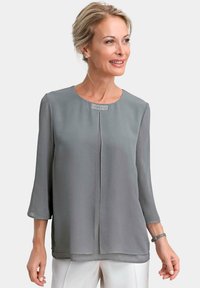 Graue Bluse mit Rundhalsausschnitt, dreiviertel Ärmeln und einer dezenten Falte. Verfügt über eine dekorative Verzierung am Ausschnitt.
