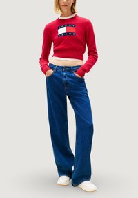 Roter kurzer Pullover mit "TOMMY JEANS" Logo, kombiniert mit weitem blauen Denim-Jeans. Modell trägt weiße Turnschuhe. Schlichter Hintergrund.