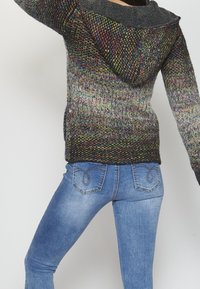 Multikleurige gebreide trui met een capuchon en lange mouwen, gecombineerd met slim-fit blauwe jeans met decoratieve stiksels op de achterzakken.