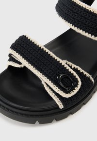 Black crochet sandals with beige detailing, featuring two adjustable straps and a textured black rubber sole. Accented with a black hardware emblem.