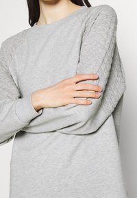 Sweatshirt gris clair avec texture matelassée sur les manches, poignets côtelés et un col rond classique. Tissu doux et confortable.