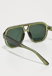 Ochelari de soare verzi cu un cadru translucid, având lentile întunecate și un design geometric minimalist. Accentuați cu un detaliu elegant al balamalei.