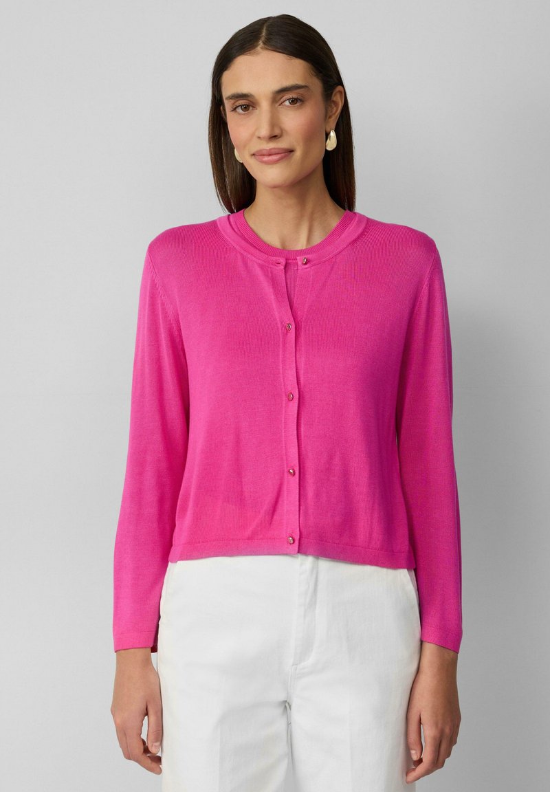 Cardigan rose léger avec un col rond, fermeture à boutons et manches longues. Texture côtelée, design court, porté sur un pantalon blanc.