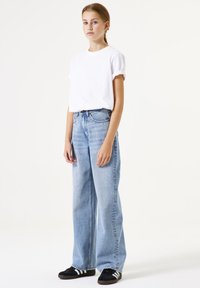 Camiseta blanca de algodón con mangas dobladas, combinada con unos vaqueros de pierna ancha de color azul claro. Calzado: zapatillas negras con rayas blancas.