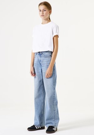 Weißes Baumwoll-T-Shirt mit aufgerollten Ärmeln, kombiniert mit hellblauen weiten Jeans. Fußbekleidung: schwarze Sneakers mit weißen Streifen.