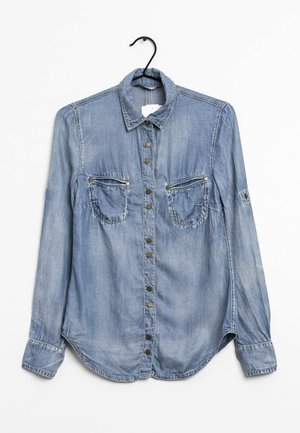 Camicia in denim di colore azzurro chiaro con abbottonatura frontale, due tasche sul petto, bottoni metallici e orlo curvo. Maniche lunghe con dettagli ai polsini.