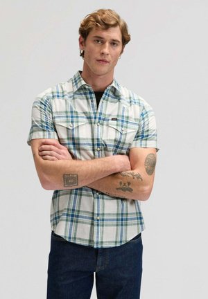 Junger Mann mit hellbraunen Haaren, der ein blau-grünes kariertes Hemd mit kurzen Ärmeln trägt und mit verschränkten Armen vor einem einfarbigen Hintergrund steht.