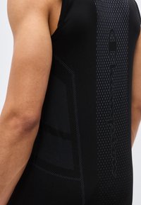 Κοντινό πλάνο του πάνω μέρους του βραχίονα και της πλάτης ενός ατόμου που φοράει μαύρο αθλητικό τοπ χωρίς μανίκια με υφασμένα μοτίβα και κατακόρυφη λεπτομέρεια λογότυπου.