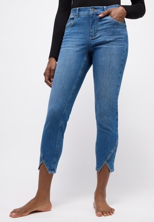 Frau trägt gekürzte blaue Jeans mit vorderen Schlitzen am Saum, steht barfuß mit einer Hand in der Tasche und trägt ein schwarzes Langarm-Top.