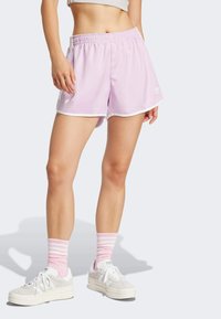 Donna che indossa dei pantaloni sportivi leggeri viola con bordi bianchi, calze a strisce rosa e bianche, e scarpe Adidas bianche, in piedi su uno sfondo bianco.