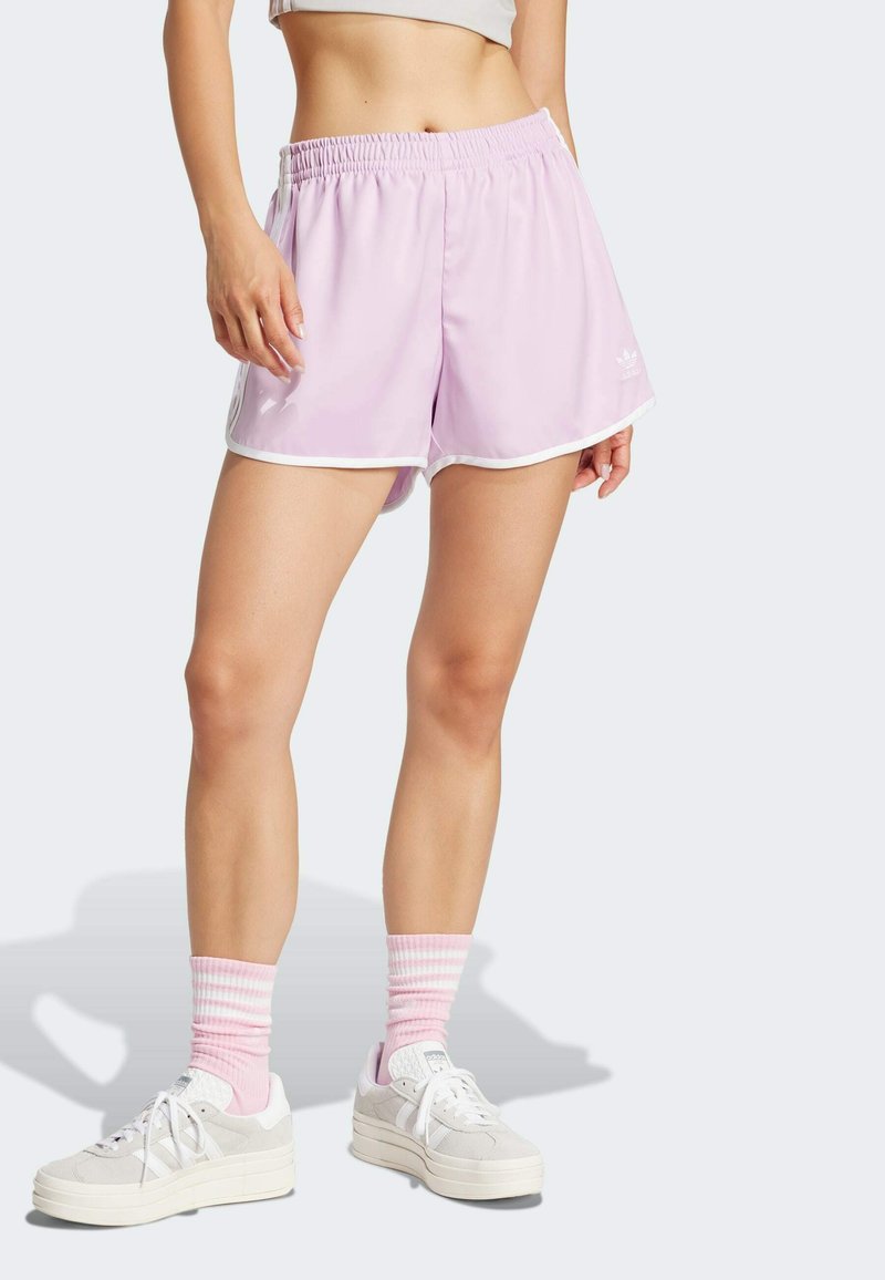 Donna che indossa dei pantaloni sportivi leggeri viola con bordi bianchi, calze a strisce rosa e bianche, e scarpe Adidas bianche, in piedi su uno sfondo bianco.