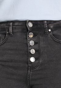 Jeans pretos de cintura alta com uma textura suave, apresentando cinco botões prateados na frente e bolsos padrão nas laterais.