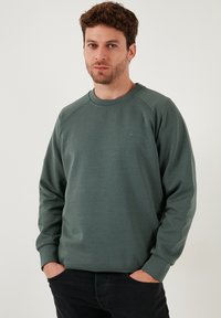 Zelená mikina vyrobená z textúrovanej látky s okrúhlym výstrihom a raglánovými rukávmi. Disponuje malým logom na hrudi. Nosená s tmavými džínsami.