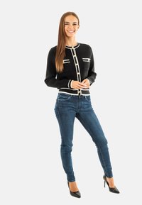 Cardigan nero a maglia con accenti bianchi, chiusura a bottoni e due tasche frontali, abbinato a jeans skinny blu scuro e tacchi alti neri.