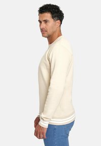 Sudadera beige con cuello redondo y puños acanalados. Presenta rayas blancas en el dobladillo y una tela texturizada. Llevada con vaqueros azules.