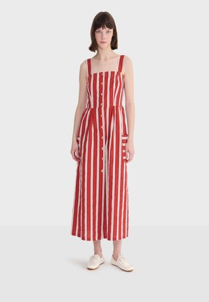 Donna che indossa un vestito midi senza maniche a righe verticali rosse e bianche con bottoni sulla parte anteriore e scarpe da ginnastica bianche su uno sfondo semplice.