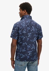 Camisa de manga corta en tejido azul marino con un patrón floral azul, que presenta un cuello puntiagudo y un corte relajado. La vista trasera muestra una textura sutil.