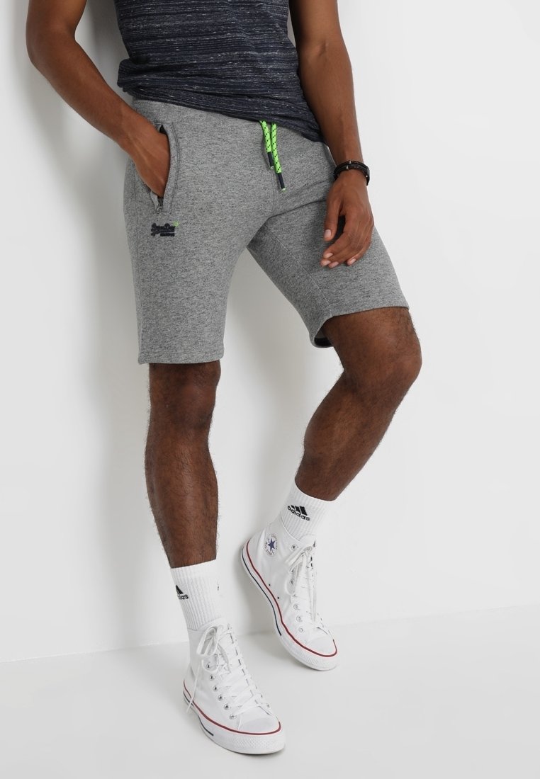 Homme portant un short gris à cordon, une chemise bleu marine, des chaussettes blanches Adidas et des baskets montantes blanches Converse, debout contre un mur blanc.
