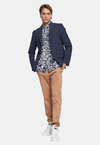 Blazer blu navy sopra una camicia floreale bianca, pantaloni beige e sneakers bianche. Il blazer presenta un motivo testurizzato e una forma strutturata.