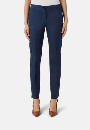 Donna che indossa pantaloni blu navy aderenti e tacchi marroni a punta, in piedi con le braccia rilassate lungo i fianchi su sfondo bianco.