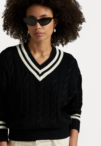 Pull en maille noire avec un col en V et des accents de rayures blanches. Présente des manches amples et un ourlet côtelé. Porté avec des lunettes de soleil noires en forme de chat.