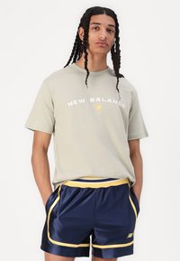 T-shirt en coton gris clair avec "NEW BALANCE" imprimé en blanc et jaune. Short marine avec des accents jaunes et des poches latérales, fabriqué en matériau brillant.