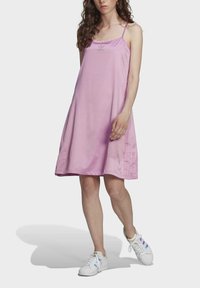 Różowa satynowa sukienka z cienkimi ramiączkami, lekko rozszerzająca się sylwetka, detale na bocznych szwach oraz haftowane logo na biuście. Połączona z białymi trampkami.
